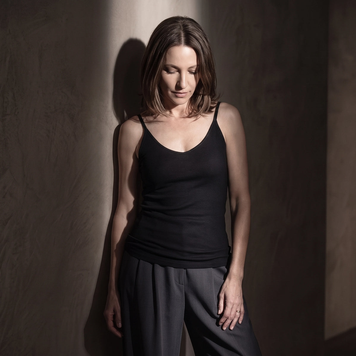 Black ribbed built-in bra camisole with dark grey pleated pants in low-key light, looking down