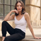 Woman seated on a stone step in a white V-neck built-in bra camisole and black jogger pants, chin resting on her hand