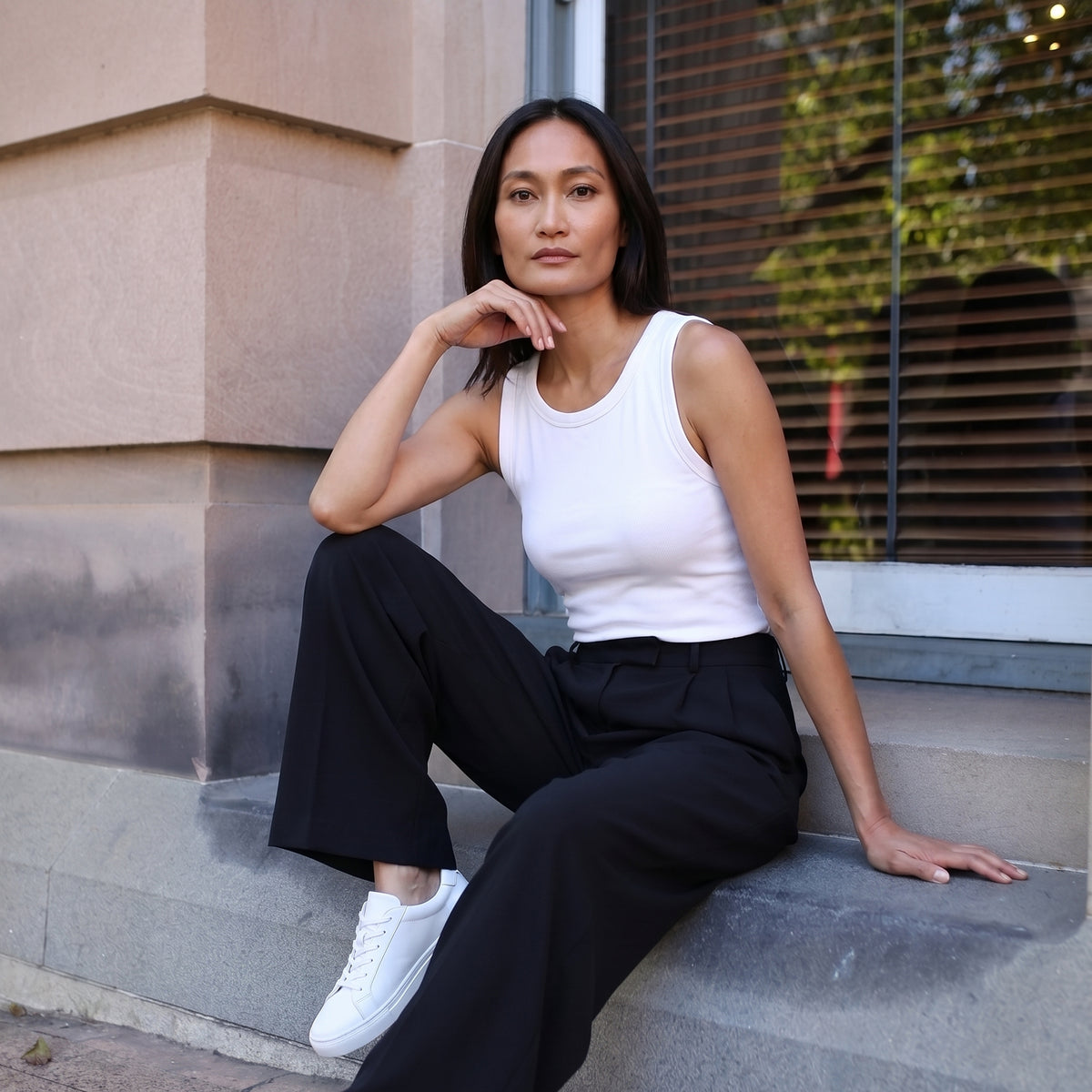 Woman sitting on steps in an off white built-in bra tank top, black pants and white sneakers