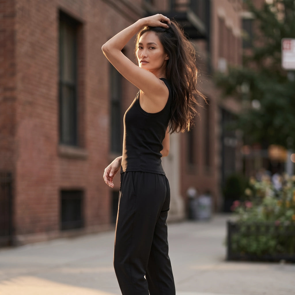 Black built-in bra tank top with black jogger pants, one arm raised to her hair in warm morning sunlight