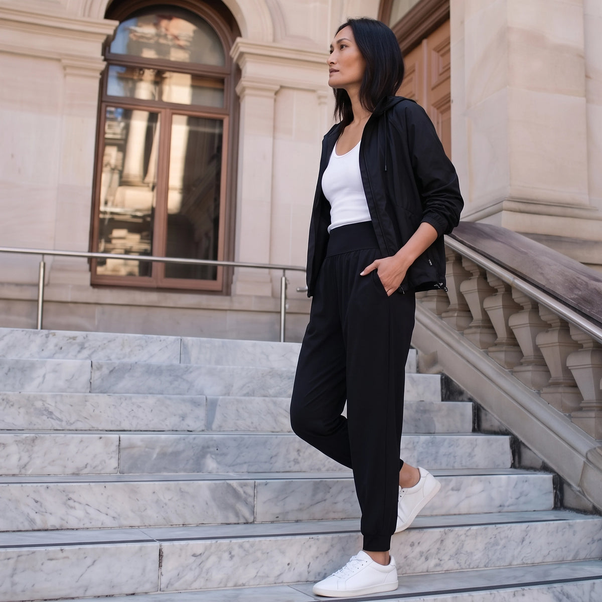 Woman in black jersey jogger pants, a black zip jacket and white sneakers walking down marble steps