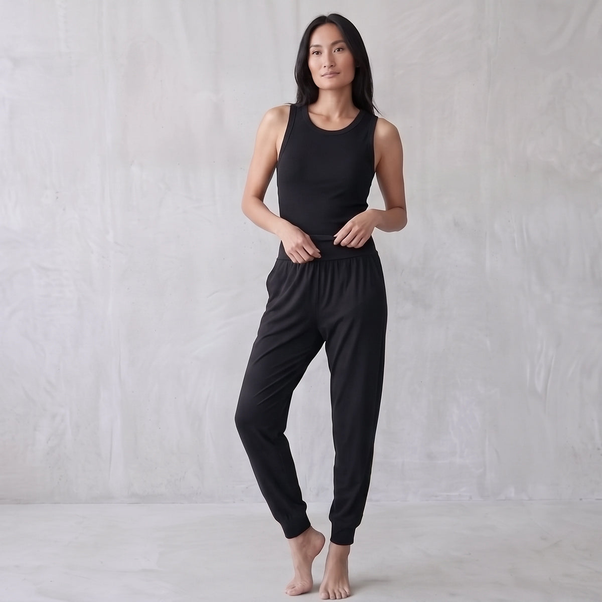 Woman in a black ribbed tank top and black jogger pants with a wide waistband, touching the waistband.