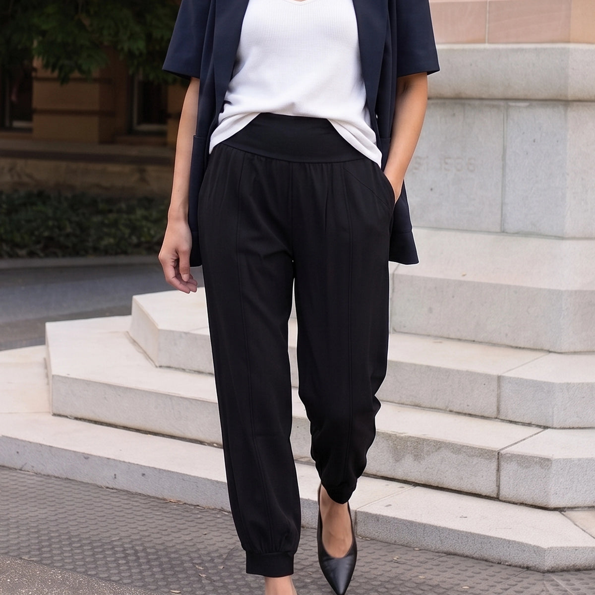 Woman in black woven jogger pants, a navy blazer and a white camisole, with one hand in her pocket