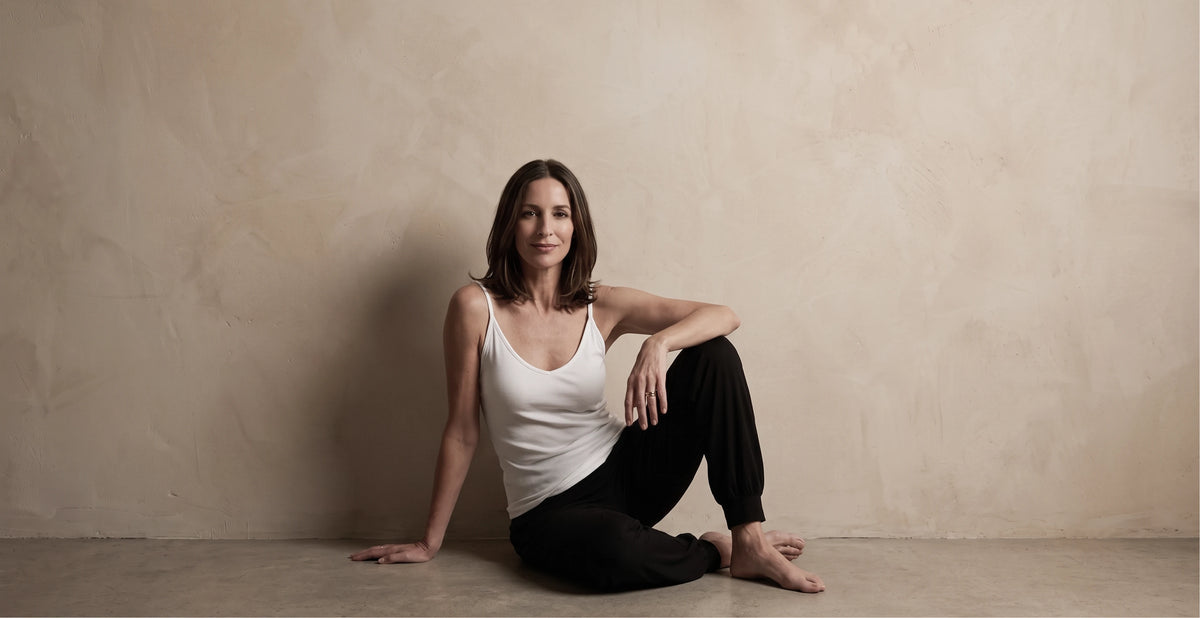 Woman sitting on the floor, resting on one arm, in a white built-in bra camisole and black jogger pants, relaxed and comfortable